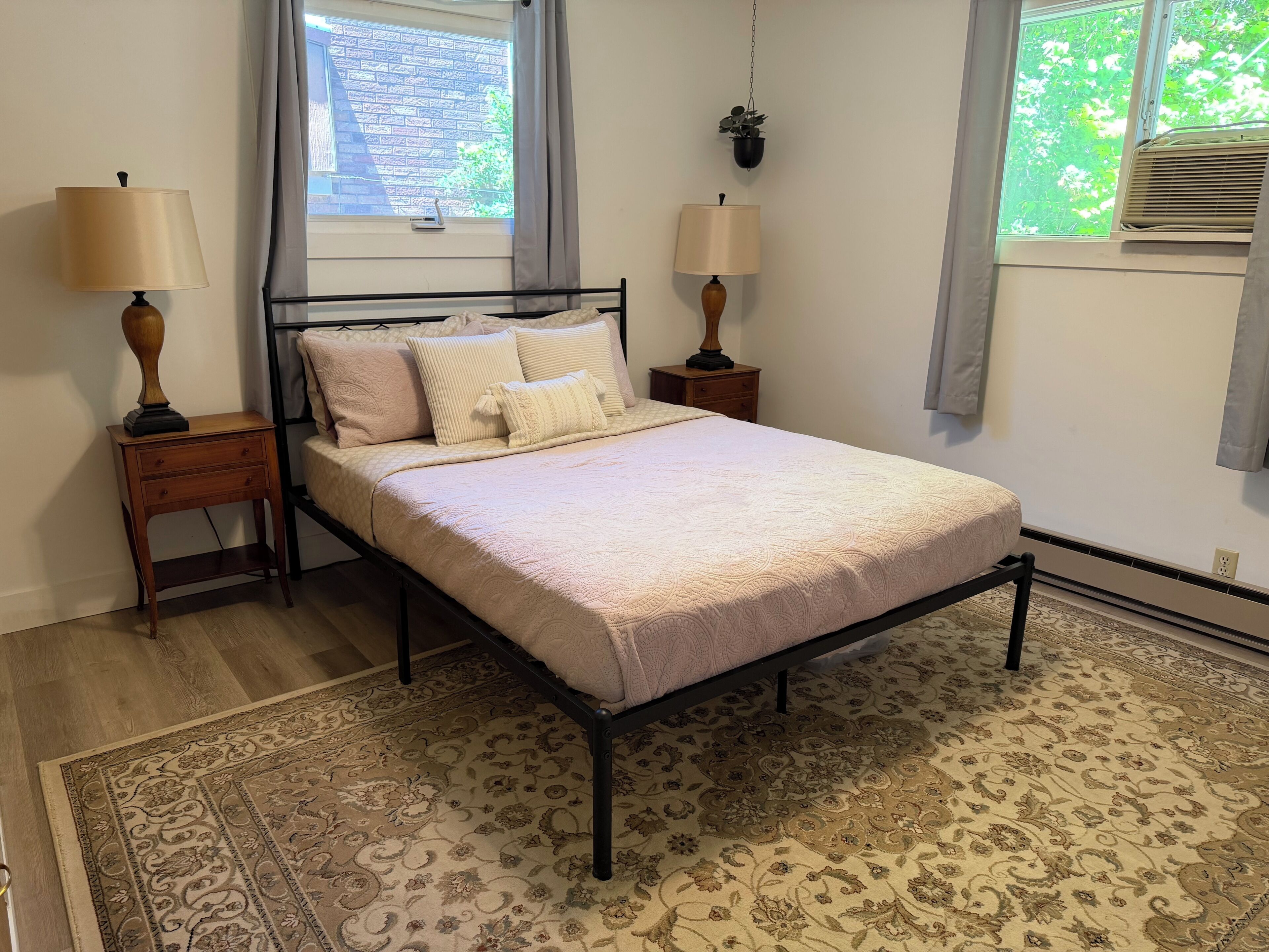 Un dormitorio ordenado con una cama, dos mesitas de noche, una ventana con cortinas, una planta colgante y una alfombra estampada.