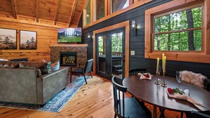 Interior acogedor de una cabaña con chimenea, paredes de madera, una mesa de comedor lista para dos personas y un ventanal con vistas a un bosque.