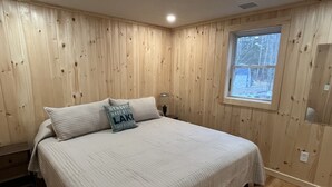 A bedroom with wooden walls, a large bed with a quilt, two pillows, a nightstand with a lamp, and a window with a view of trees.