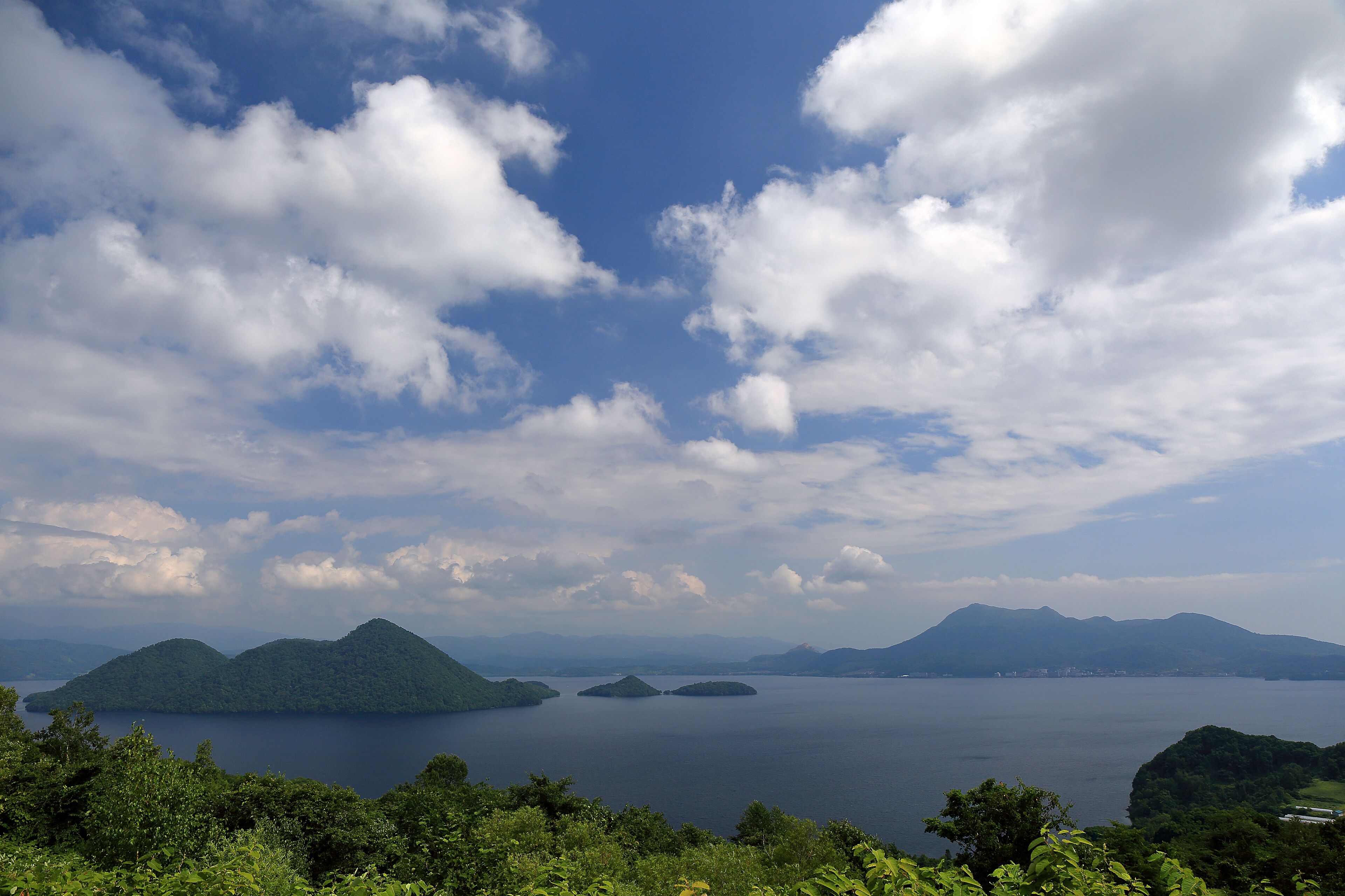 A panoramic view of a lake surrounded by lush green hills and mountains under a partly cloudy sky.