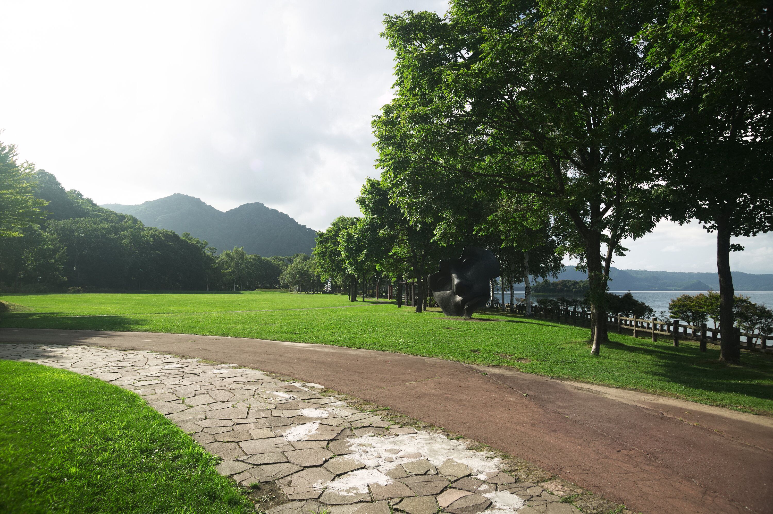 A paved pathway leading to a grassy area with trees and a view of mountains.