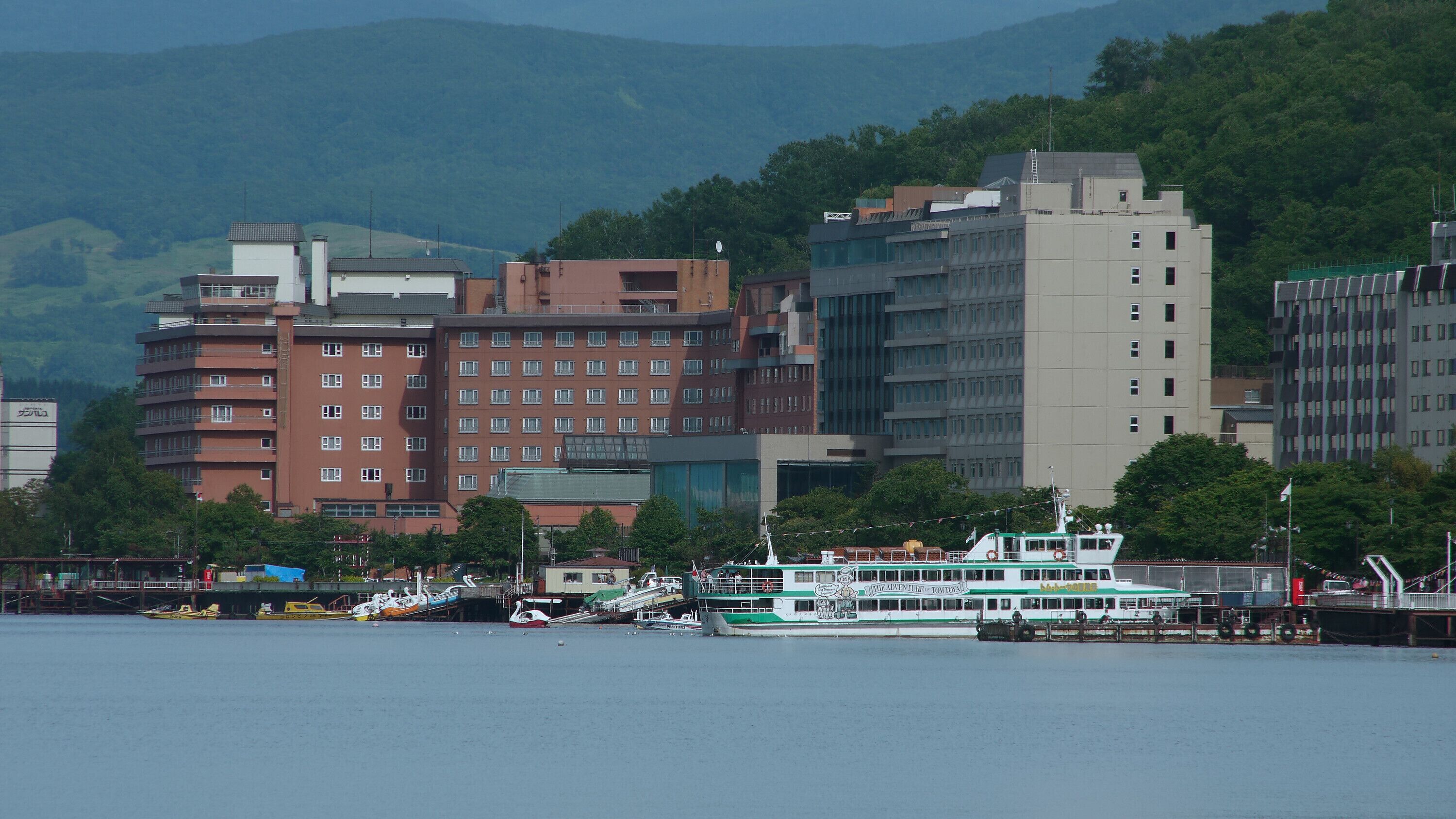 A waterfront cityscape with a large white and green boat docked at a pier, surrounded by multi-story buildings and lush greenery.