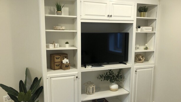 Un mobile contenitore bianco con una televisione a schermo piatto, mensole decorate con oggetti ornamentali e una pianta in vaso.