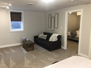 Una habitación pequeña y bien iluminada con un sofá de color oscuro, una mesita de madera y una ventana con persianas. Encima del sofá hay tres adornos en la pared y una puerta que conduce a otra habitación.