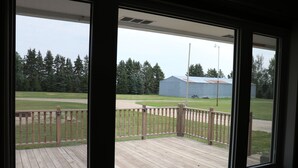 A view from a window overlooking a wooden deck with a railing, a grassy field, and a building in the distance.