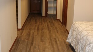 A narrow hallway with wooden flooring, a door at the end, and a bed with a patterned bedspread on the right.