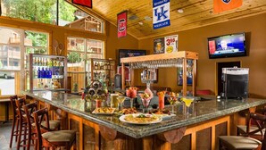 Un espace bar avec un comptoir en bois, des tabourets et une variété de boissons. On y trouve des drapeaux des Kentucky Wildcats et un écran de télévision fixé au mur.