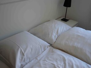 Un lit simple, sans fioritures, avec du linge de lit blanc et une seule table de chevet sur laquelle se trouve une lampe.