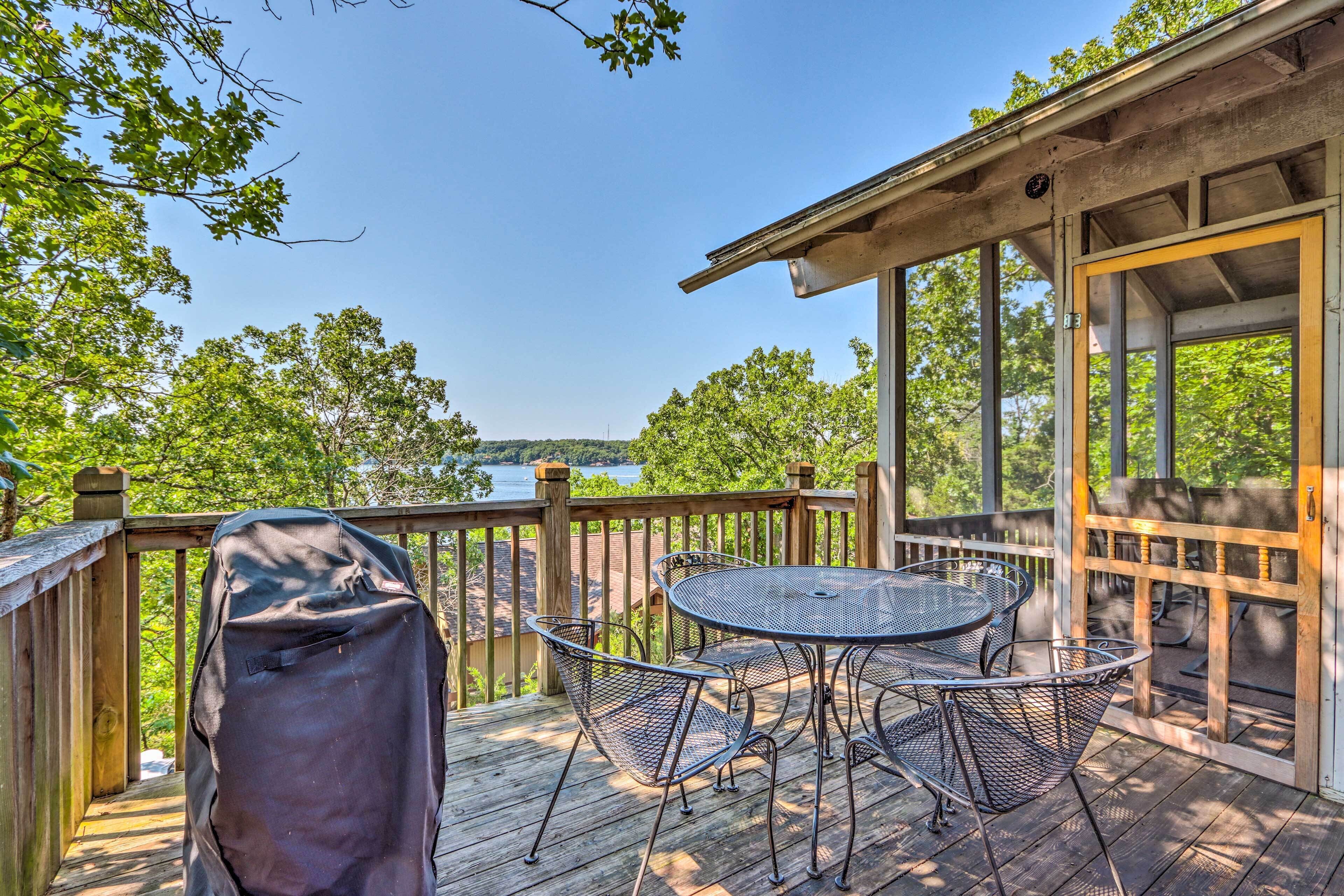 A covered outdoor deck with a metal table and chairs, overlooking a body of water and surrounded by trees.