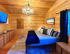 A log cabin room with a bed, a bench, a TV, and a painting on the wall.