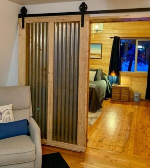 A room with a sliding door, a bed with a green blanket, a lamp, and a window with a view of trees.