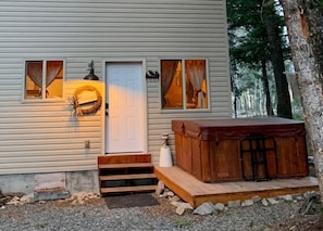 Une cabane avec une façade en bois, une porte d’entrée, un porche avec des marches, un jacuzzi et une guirlande accrochée à la porte.