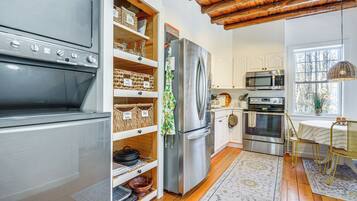 A modern kitchen with a stainless steel refrigerator, oven, and microwave. There are wooden shelves with baskets and a woven chair.