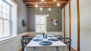 A dining room with a table set for six, a chandelier, a painting, and a window with a view of trees.