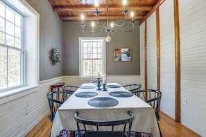 A dining room with a table set for six, a chandelier, a painting, and a window with a view of trees.