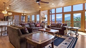 A spacious living room with a large sofa set, a coffee table, and a dining table. There is a ceiling fan and large windows offering a view of the mountains.