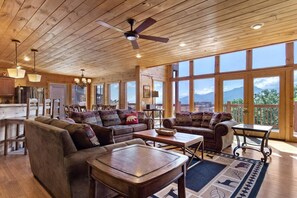 A spacious living room with a large sofa set, a coffee table, and a dining table. There is a ceiling fan and large windows offering a view of the mountains.