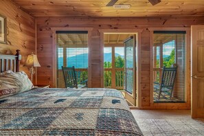 A wooden cabin bedroom with a large bed, a nightstand, a lamp, and a view of the outdoors through a sliding door.