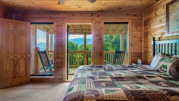 A bedroom with a large bed, wooden walls, a ceiling fan, and a view of greenery through the windows.