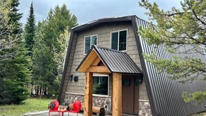A modern, two-story cabin with a sloped roof, a stone foundation, and a wooden porch with red chairs. - Trailer Parking *Dog Friendly* Fireplace (Island Park)
