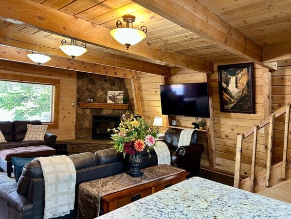 A wooden cabin interior with a stone fireplace, leather sofas, a flat-screen TV, and a staircase.