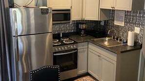 Cozinha compacta com geladeira de aço inoxidável, micro-ondas, forno e pia. Há uma mesa de madeira e um ventilador de teto.