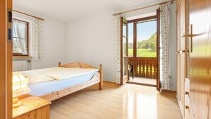 Une pièce avec un lit, une fenêtre avec des rideaux, un bureau en bois et une vue sur un paysage verdoyant, visible à travers une porte ouverte.