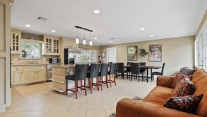 A spacious living area with a brown leather sofa, a dining table with chairs, and a kitchen area with cabinets and a refrigerator.
