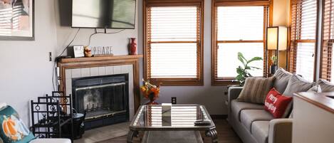 A living room with a fireplace, a sofa, a coffee table, and two windows with blinds.