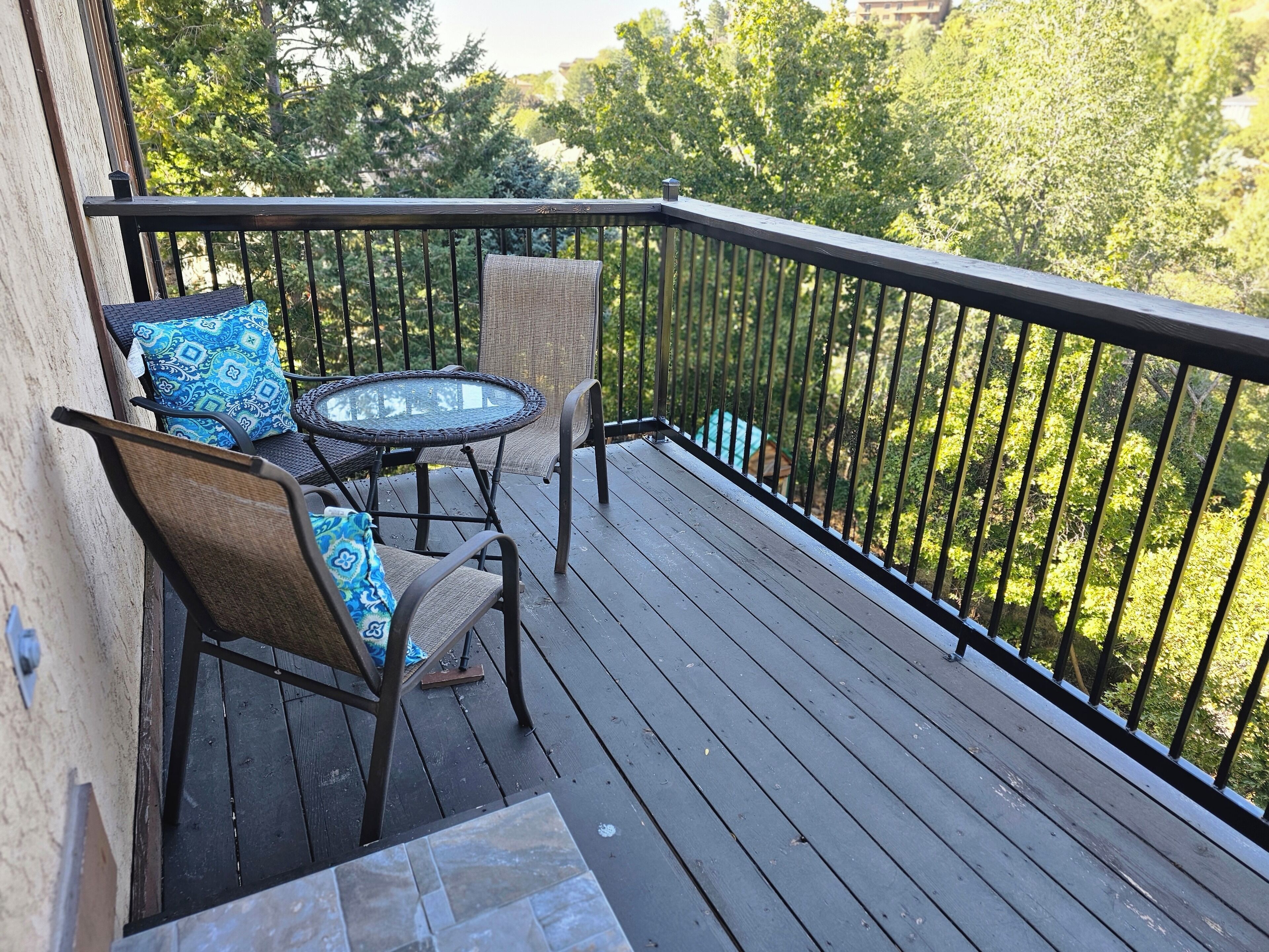 A wooden deck with a round table and two chairs, a black metal railing, and a view of trees.