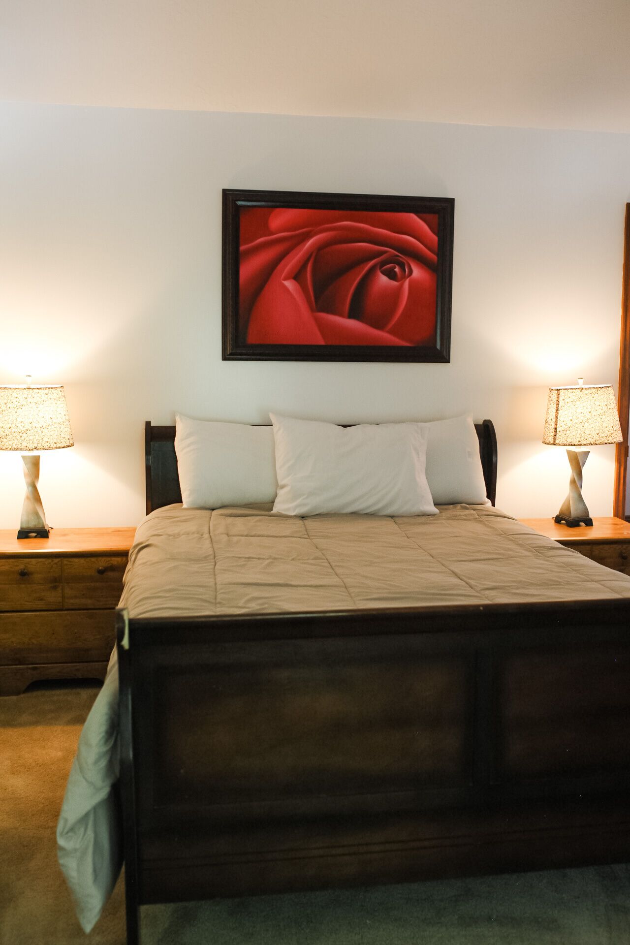 A neatly made bed with a dark headboard, two bedside tables with lamps, and a large framed picture of a red rose on the wall.