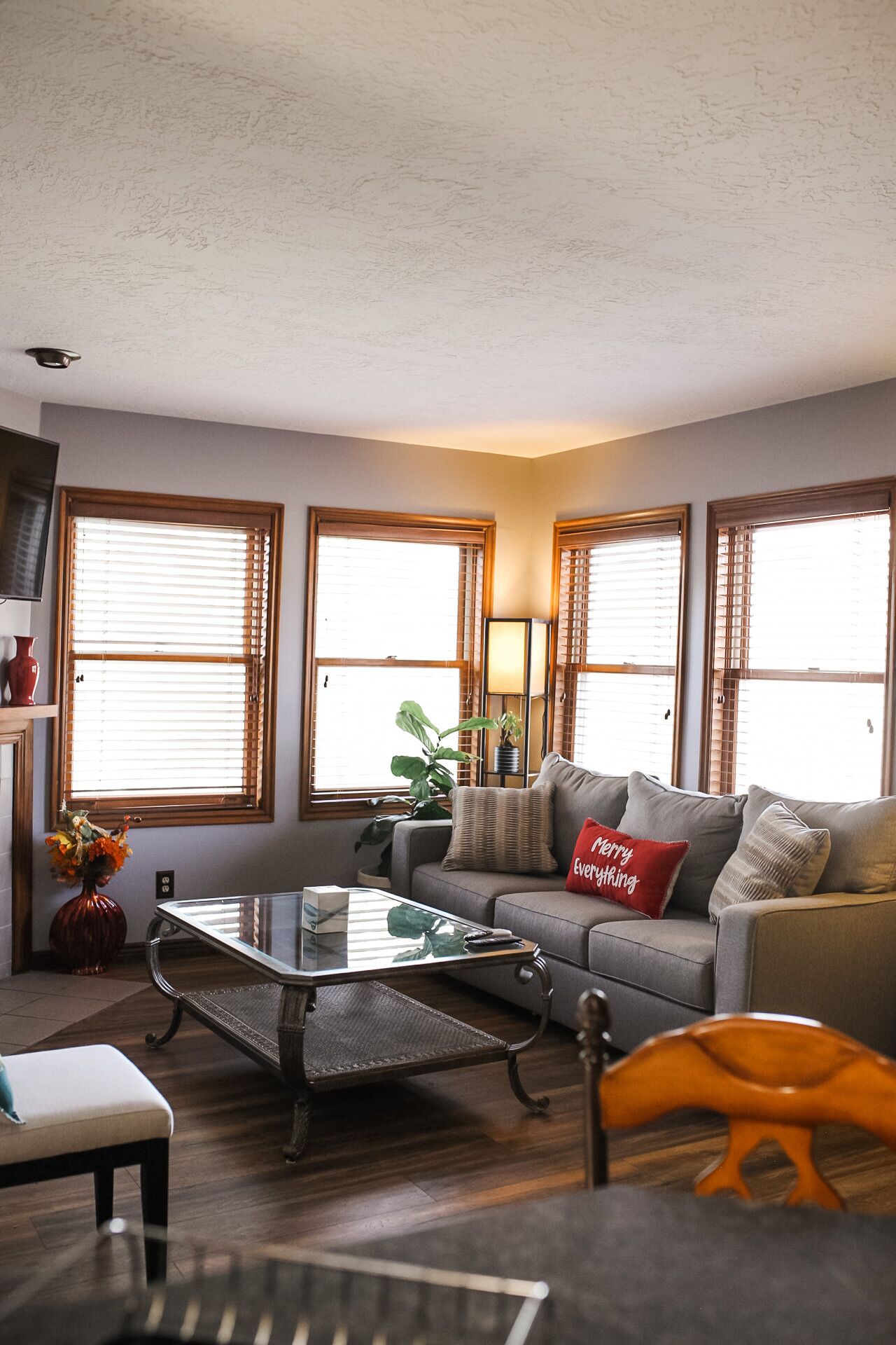 A living room with a sofa, a glass coffee table, and a wooden chair. There are three windows with blinds, a lamp, and a vase with flowers.