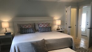 Un dormitorio ordenado con una cama grande, dos mesitas de noche con lámparas, un reloj despertador y una puerta que da a otra habitación.