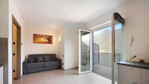 Un apartamento moderno con un sofá gris, un cuadro de un árbol, una puerta corrediza de vidrio a un balcón y una encimera de cocina.