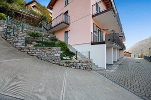A multi-story building with a stone staircase leading to a balcony, a paved walkway, and a mountainous backdrop. - Appartamento 4 Il Tornante with Lake & Mountain Views, Wi-Fi, A/C & Balcony (Nesso)