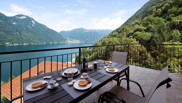 A table set with breakfast items, a view of a lake, and mountains in the background. - Appartamento 5 Il Tornante with Lake & Mountain Views, Wi-Fi, A/C & Balcony (Nesso)