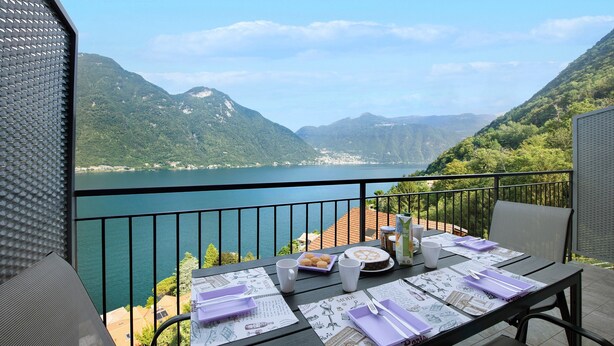 A balcony with a table set for dining, overlooking a lake and mountains. - Appartamento "7 Il Tornante" with Lake & Mountain Views, Wi-Fi, A/C & Balcony (Nesso)