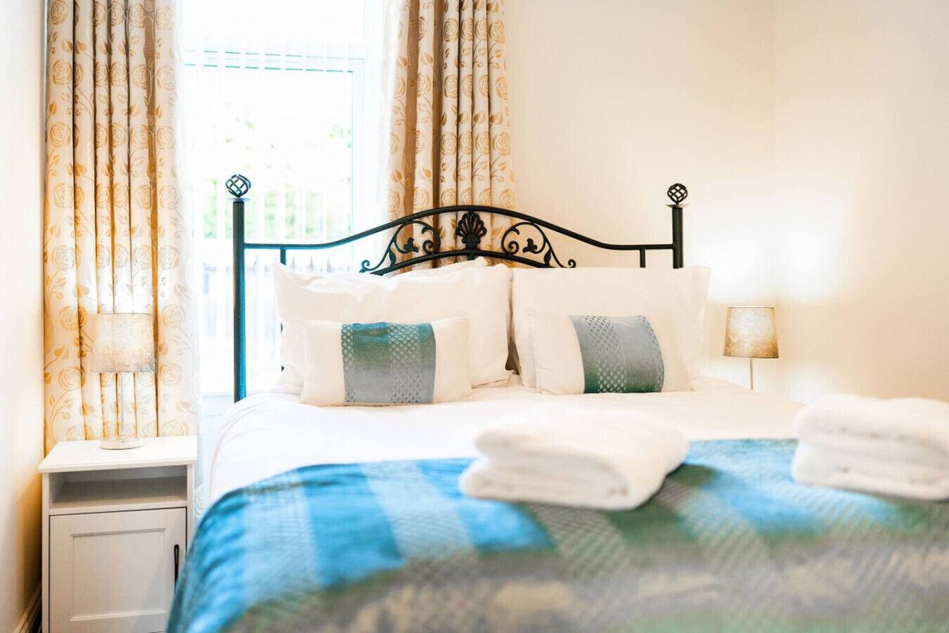 Un letto rifatto con una coperta a quadri blu e bianchi, cuscini bianchi e due asciugamani piegati sul letto. Un comodino con una lampada e una finestra con tende.