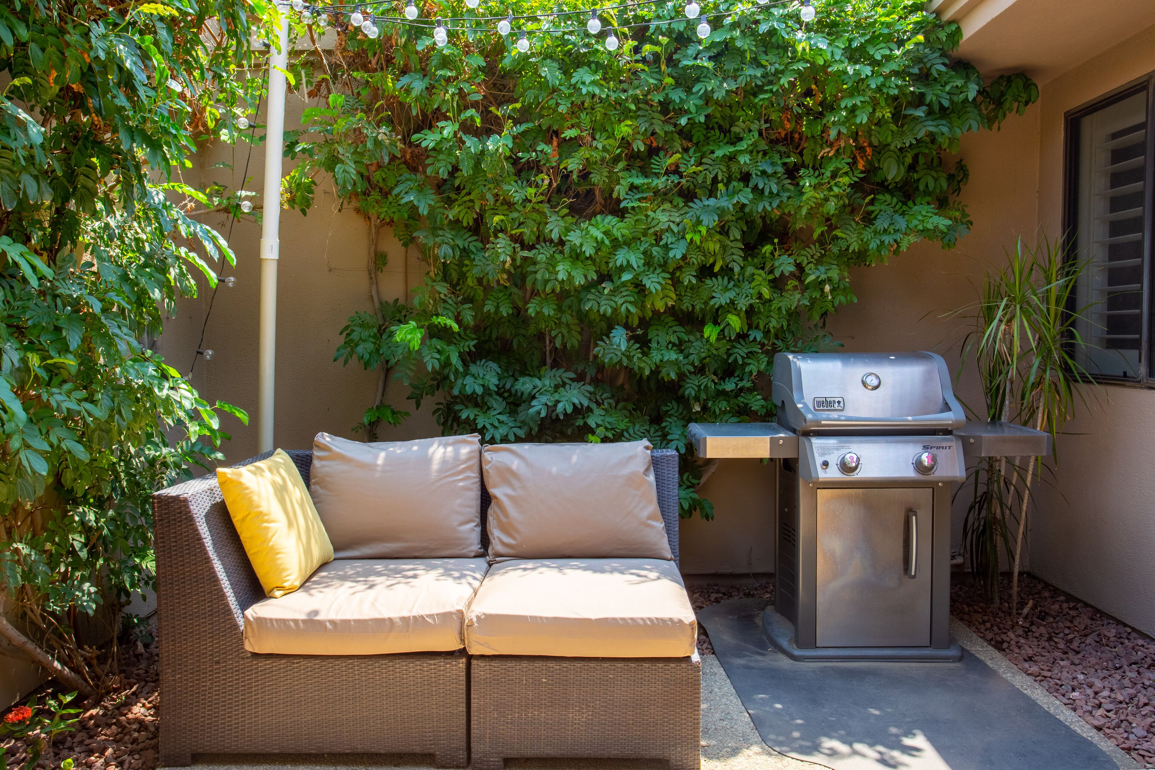 A patio with a wicker sofa, a grill, and lush greenery.
