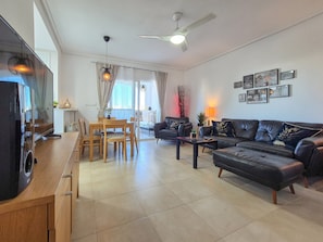 A living room with a sofa, coffee table, TV, dining table, chairs, ceiling fan, and artwork on the wall.