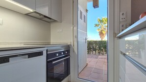 A modern kitchen with a dishwasher, oven, and a sliding glass door leading to an outdoor patio with a palm tree and a hedge. - Townhouse overlooking golf course (La Torre Golf Resort)