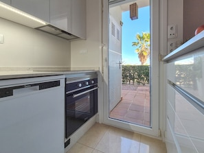 Eine moderne Küche mit Geschirrspüler, Backofen und einer Glastür, die auf eine Terrasse im Freien mit einer Palme und einer Hecke führt.