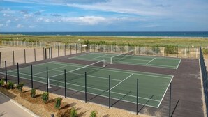Un court de tennis en plein air, avec un filet au milieu, entouré d’une clôture, et offrant une vue sur l’océan en arrière-plan.