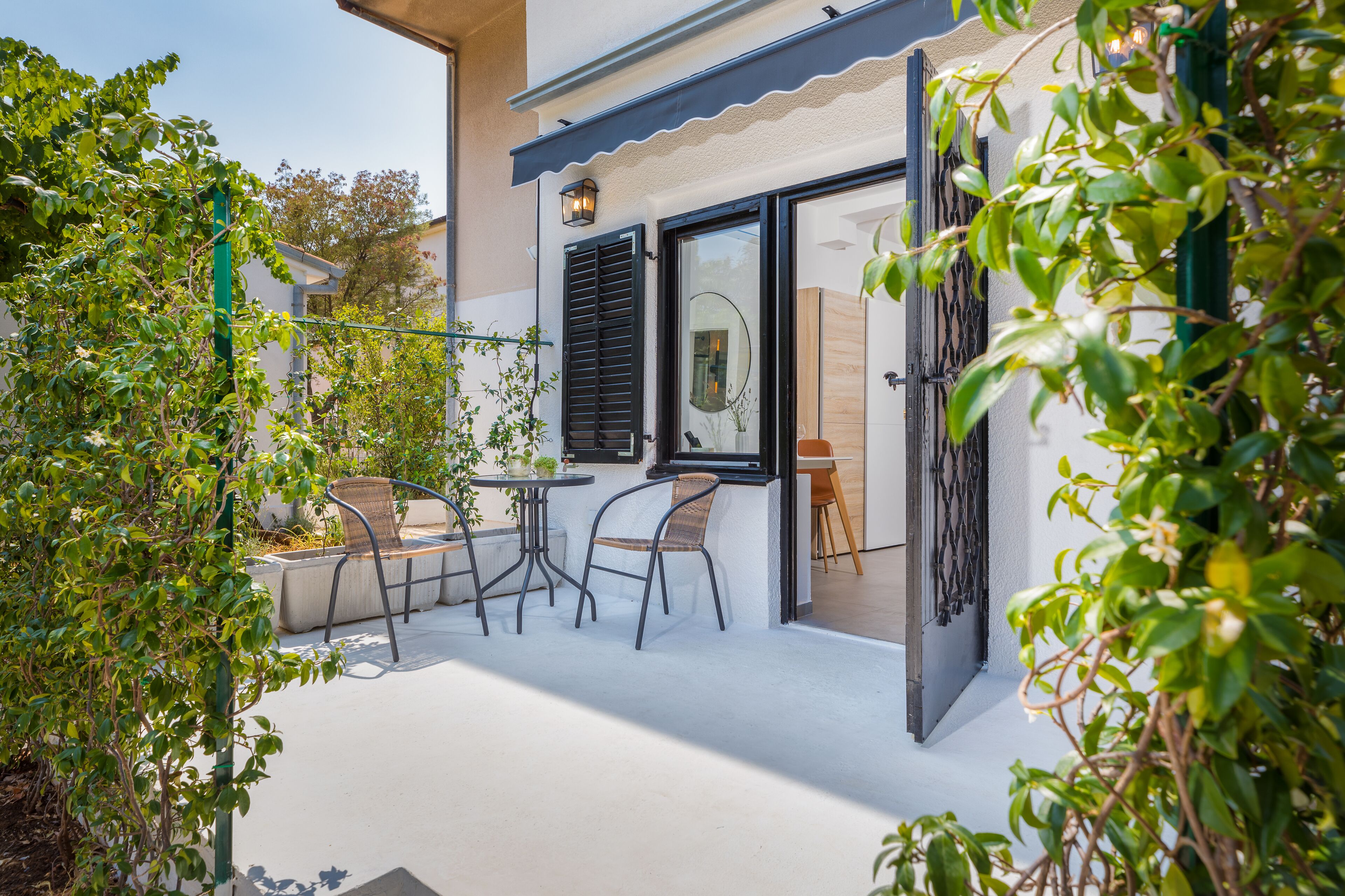 An outdoor patio with a table and chairs, a door leading to the interior, and surrounding greenery.