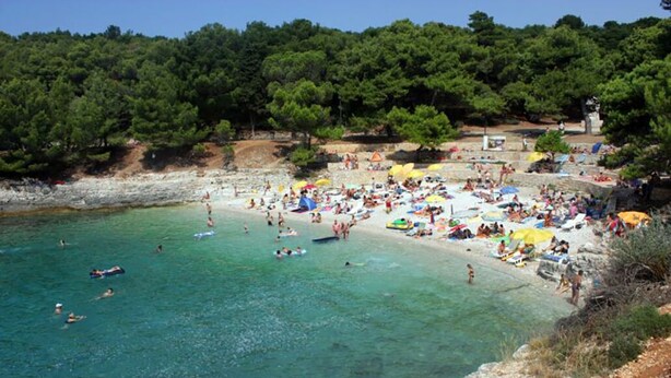 Une plage bondée, avec une eau turquoise et limpide, où l’on peut voir des gens nager et bronzer, le tout sur fond d’une végétation luxuriante.