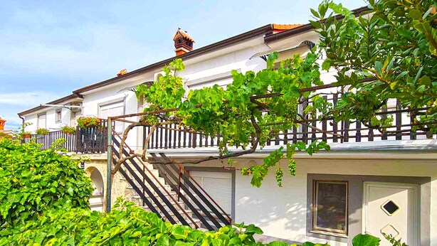 Ein weißes Gebäude mit Balkon, Treppe und einem Garten mit grünen Pflanzen.