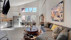 A modern living room with a grey sofa, a round coffee table, and a cowhide rug. There is a dining area with a white table and chairs, and a kitchen with white cabinets and a built-in oven.