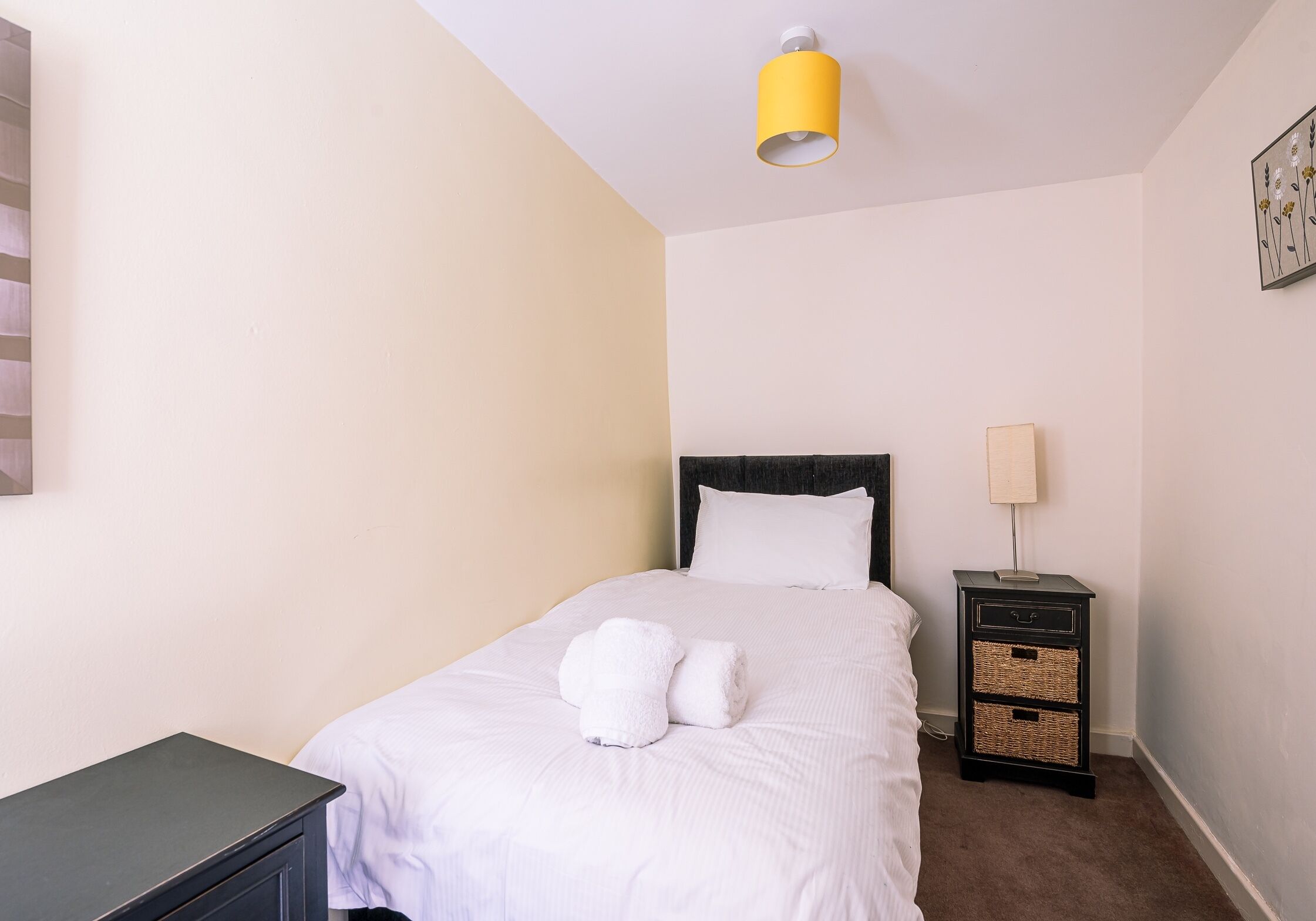 Ein ordentlich gemachtes Bett mit weißen Leinen und zwei aufgerollten Handtüchern, eine Nachttischlampe und ein kleiner schwarzer Nachttisch mit Schubladen.