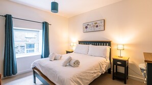 A neatly made bed with white linens and rolled towels, a bedside table with a lamp, and a window with curtains. - Beautiful, 2 bedroom, first floor apartment, at the heart of a busting Dales Village. (Richmond)