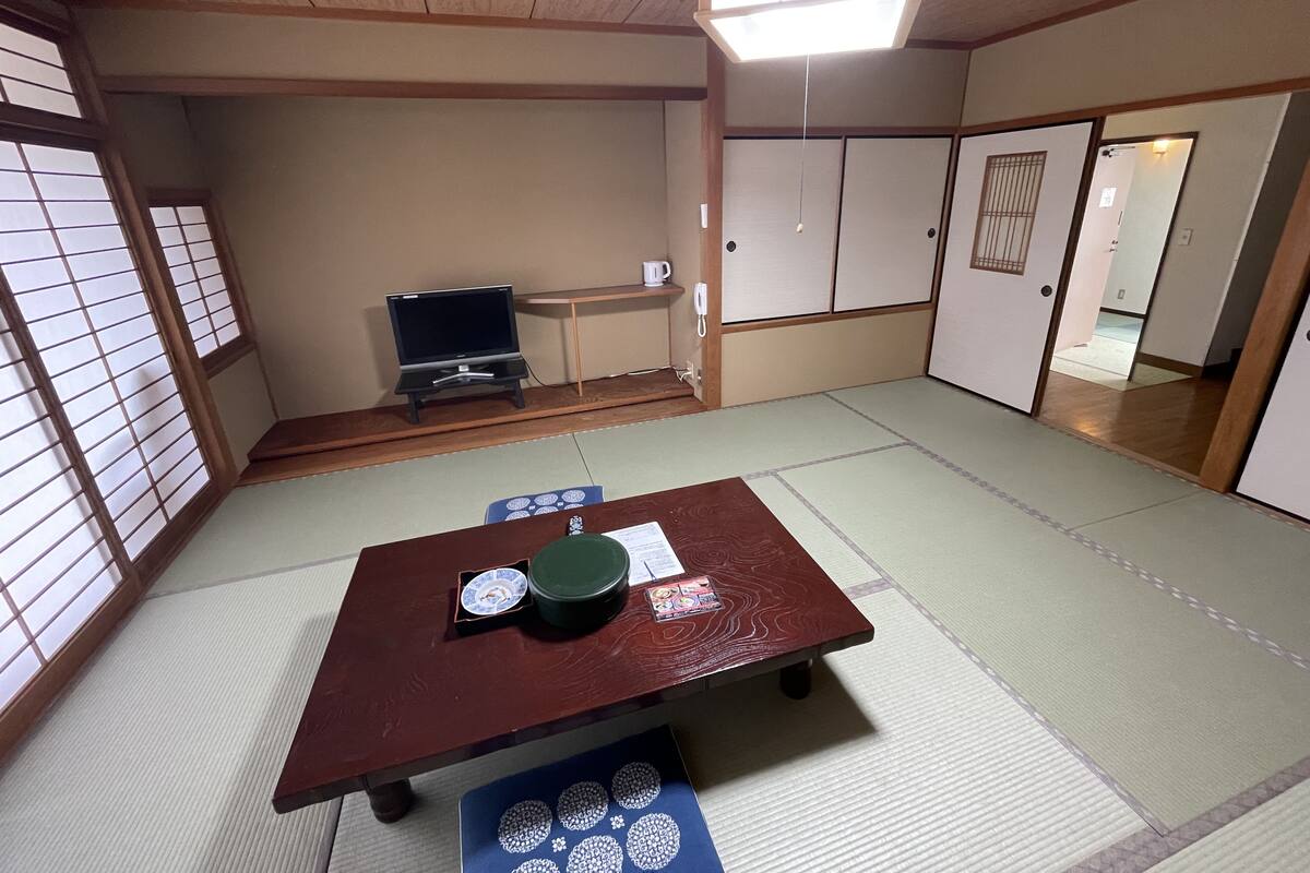 Une pièce de style japonais traditionnel, avec un sol recouvert de tatamis, une petite table sur laquelle repose un plateau vert, un téléviseur posé sur un meuble et des portes coulissantes.